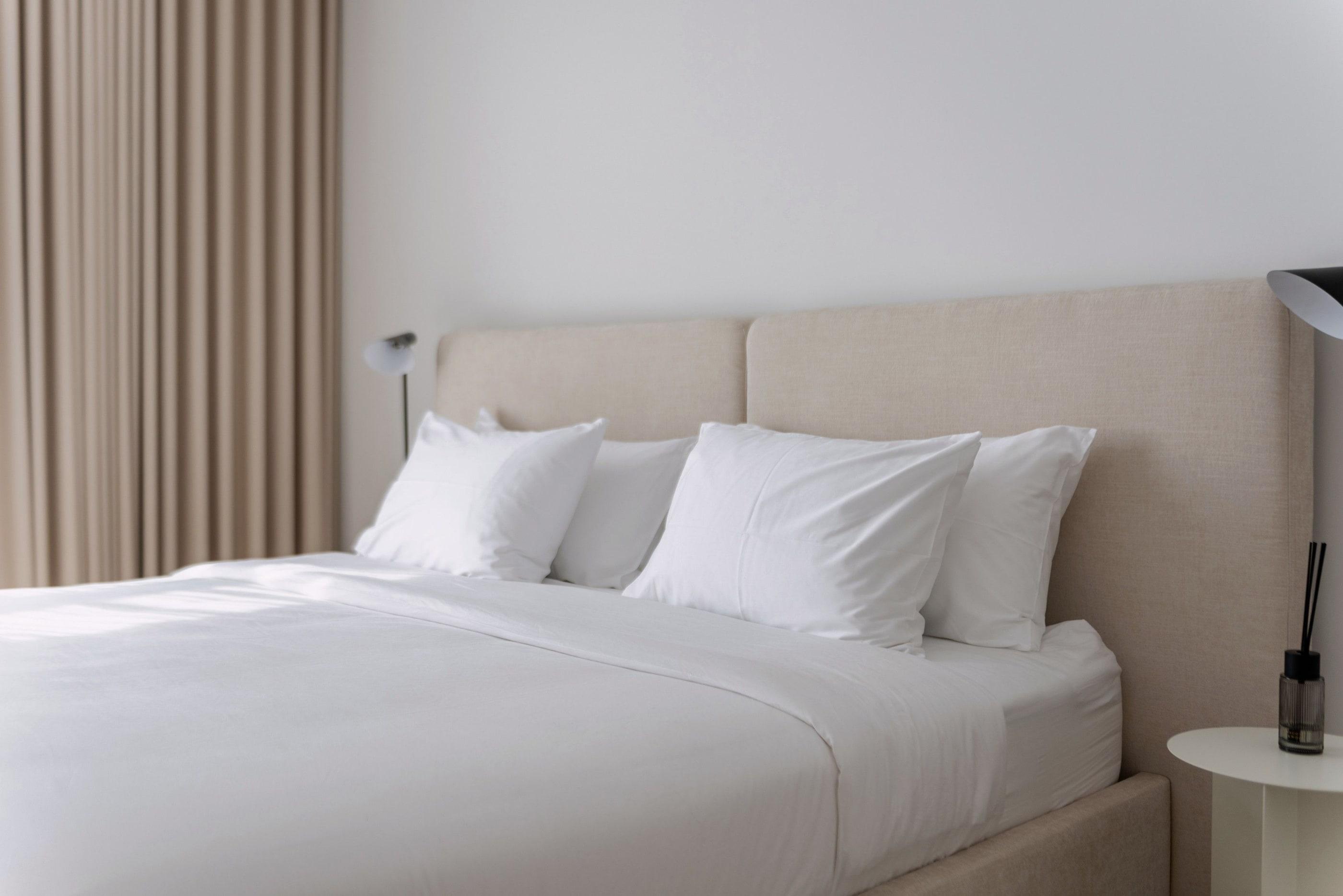 Neutral bedroom with neatly made white bedding, an upholstered headboard and soft curtains